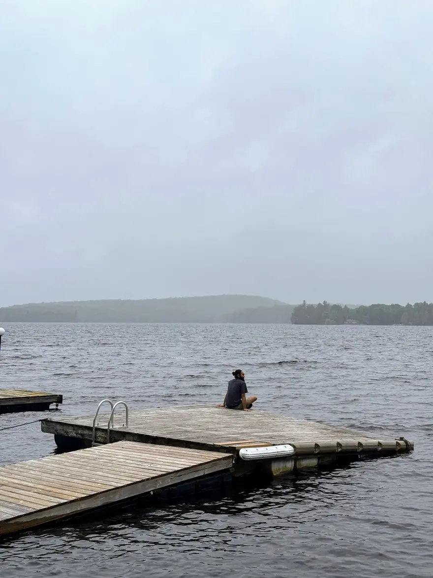 Harsimran sitting alone on a wooden dock, looking out over a calm grey lake